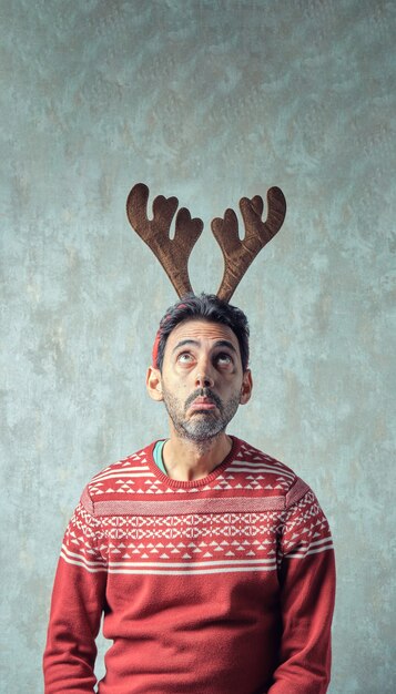 Dark boy with short gray beard in red christmas sweater and headband of reindeer antlers looking up with grimace on face