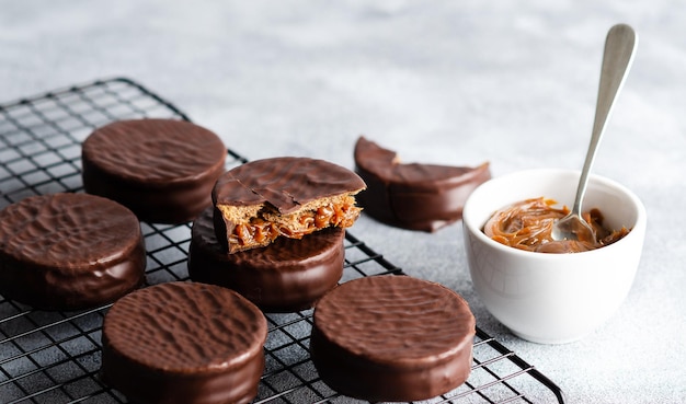 Dark chocolate alfajores on a dark rack and a bowl with dulce de leche, on grey background.