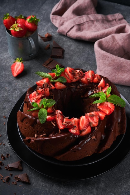 Dark chocolate bundt cake with ganache icing and strawberry on\
dark stone or concrete table background festive cake selective\
focus