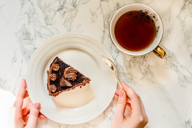 休日のチョコレート釉薬とお茶のゴールドカップとダークチョコレートケーキ。甘い朝食。若い女性の手。食べる過程。上面図。