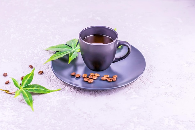 香りのよいコーヒーとコナビスの葉が皿の上にある暗いカップは、灰色のコンクリートの背景の上に立っています。エネルギーのためのモーニングコーヒーの概念。
