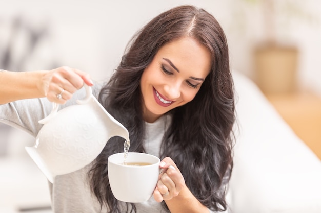 Dark curly haired woman pouring hot coffee or tea into white cup.