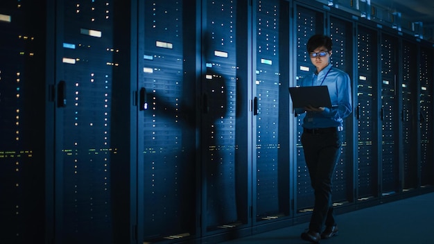 In Dark Data Center Male IT Specialist Walks along the Row of Operational Server Racks Uses Laptop