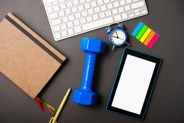 A dark desk with a planner, a blue weight, a tablet, a keyboard for making the time more productive