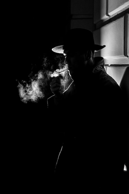 Dark dramatic silhouette of a man in a hat Smoking a cigarette on the street at night in Noir style