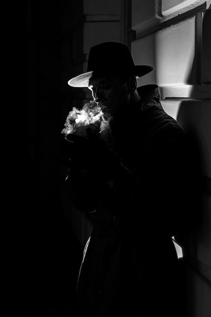 Dark dramatic silhouette of a man in a hat Smoking a cigarette on the street at night in Noir style