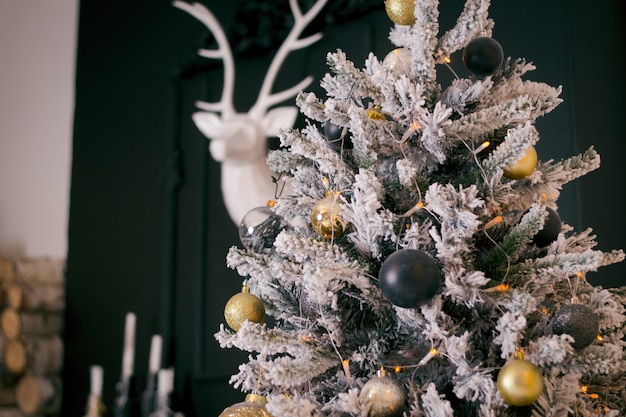 그것에 흰색 인공 눈이 어두운 녹색 크리스마스 트리. 금색과 진한 파란색 장신구와 고딕 크리스마스 트리. 벽에 흰 사슴과 어두운 인테리어입니다. 새해의 개념