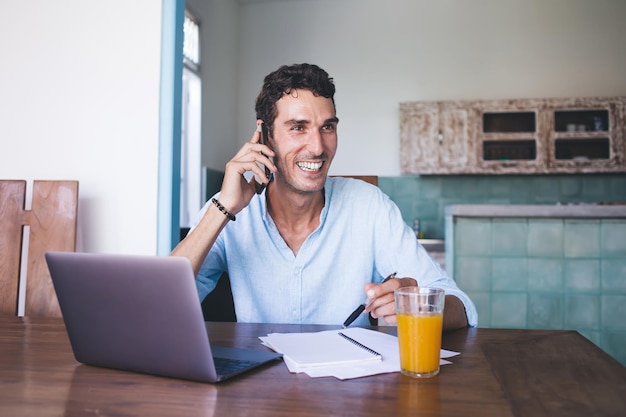 Dark haired adult male in casual clothes looking away and laughing while sitting at laptop with notepad and orange drink and making phone call in light apartment