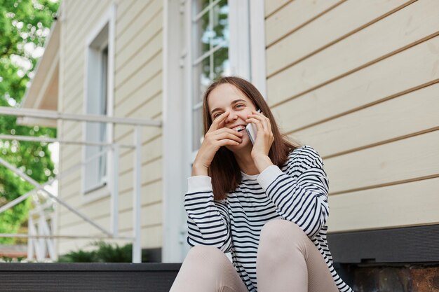 Dark haired adult woman wearing striped shirt and leggins sitting on house porch talking on mobile phone smiling happily having conversation with friends