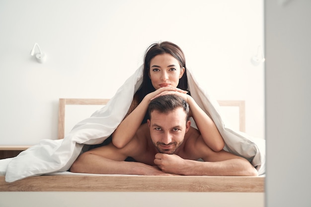 Темноволосая привлекательная молодая женщина под одеялом, опираясь руками на голову мужа