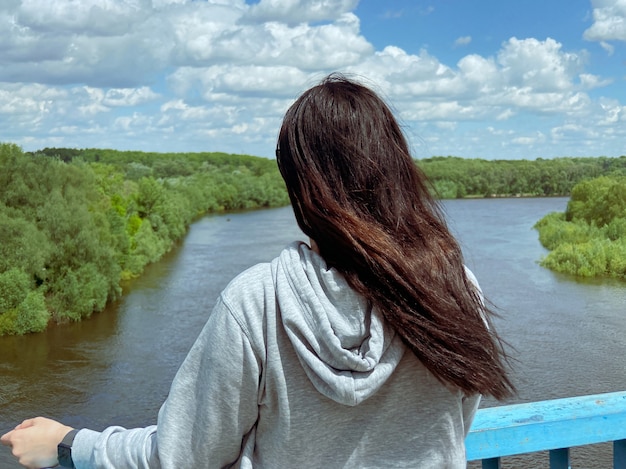 A dark haired Ñaucasian girl stands with her back in a light gray jacket and looks towards the river and the forest in the city of Chernigov in Ukraine.