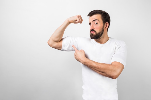dark-haired and bearded guy standing near white wall and pointing at his big muscles on his right hand and looking to camera. This guy is confident he has good body shape.