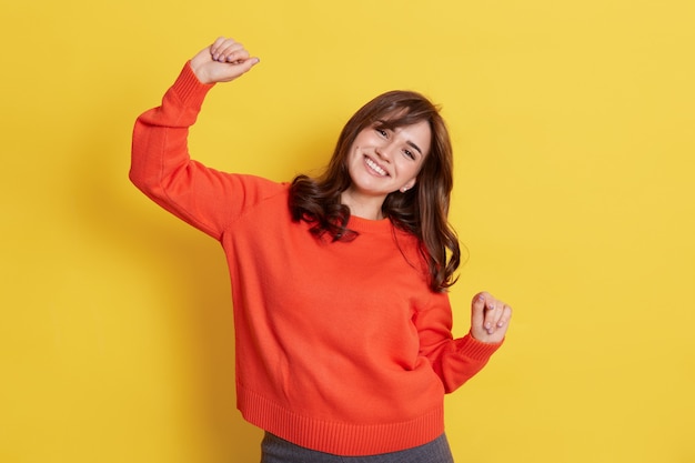 Dark haired beautiful young woman model in casual clothing dancing happily, celebrating pleasant event, raising hands.