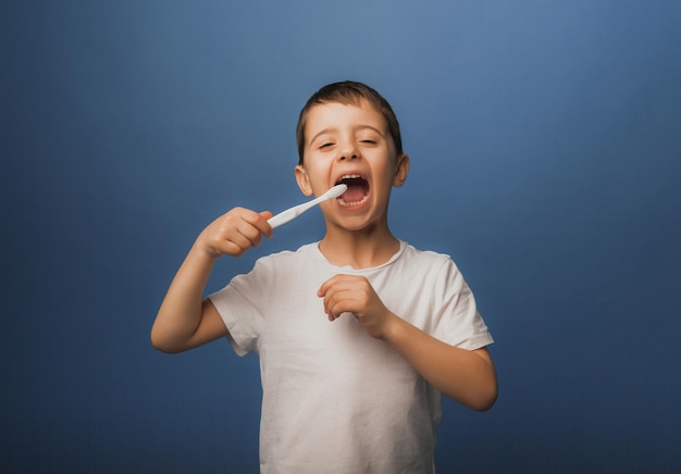 A dark-haired boy in a white T-shirt on a blue background brushes his teeth with a toothbrush. baby hygiene