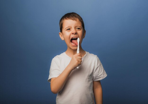 A dark-haired boy in a white T-shirt on a blue background brushes his teeth with a toothbrush. baby hygiene