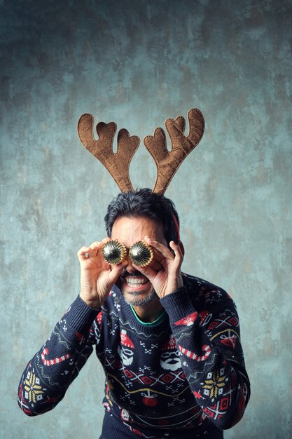 Photo dark-haired boy with a short gray beard in a blue christmas sweater and a reindeer antler headband holds two golden christmas baubles in front of his eyes