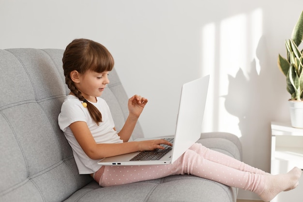 Dark haired cute adorable little girl with pigtail sitting on
sofa in light living room with laptop on her knees, typing on
keyboard, choosing cartoon for watching.