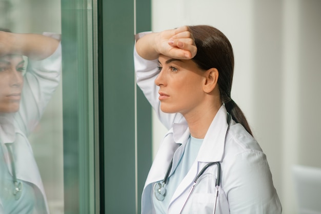 A dark-haired doctor standing near the window and looking thoughtful