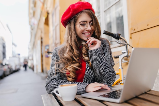 Studentessa dai capelli scuri con occhiali alla moda che lavora con il computer portatile durante il pranzo al bar. ritratto all'aperto di un romantico freelance in berretto rosso che utilizza il computer e si gode un caffè caldo.