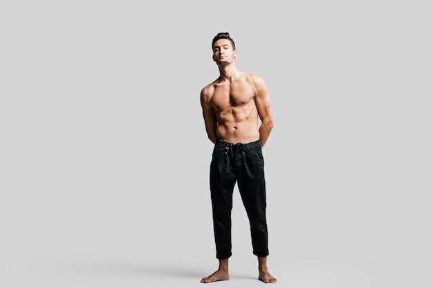 Dark-haired handsome young dancer with bare torso wearing a black sports pants is standing on a white background