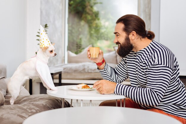 Dark-haired man feeling relaxed and rested while drinking latte and looking at his pet
