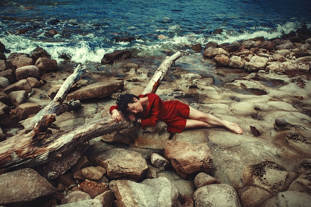 Dark haired model is posing in a red dress on the sea bank