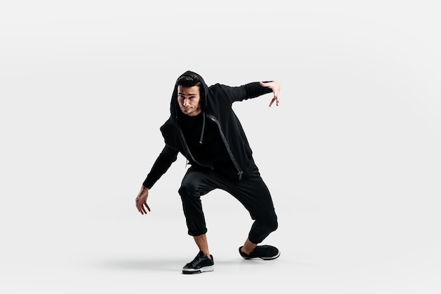 Темноволосый стильный молодой человек в черной толстовке и черных брюках делает стилизованные движения уличных танцев.