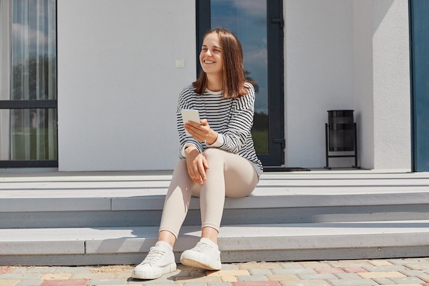 Dark haired young adult woman wearing striped shirt using phone posing near house sitting on porch holding smartphone looking away checking social networks