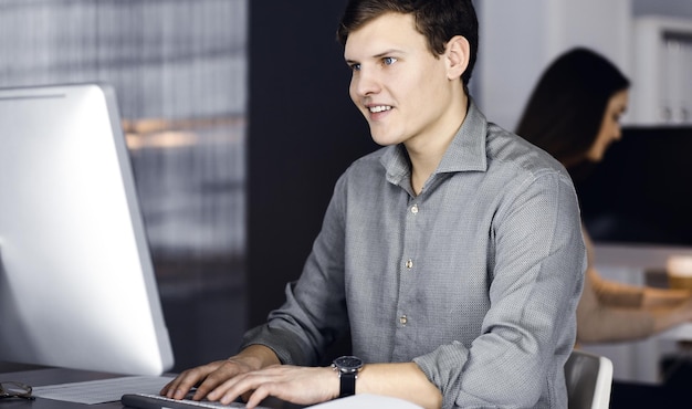 녹색 셔츠를 입은 검은 머리의 젊은 사업가이자 프로그래머가 컴퓨터 작업을 열심히 하고 있으며 현대적인 캐비닛의 책상에 앉아 있고 배경에는 여성 동료가 있습니다. 성공의 개념