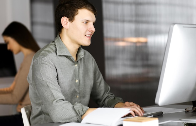 Photo dark-haired young businessman and programmer in a green shirt is working hard on his computer, while sitting at the desk in a modern cabinet with a female colleague on the background. concept of succe