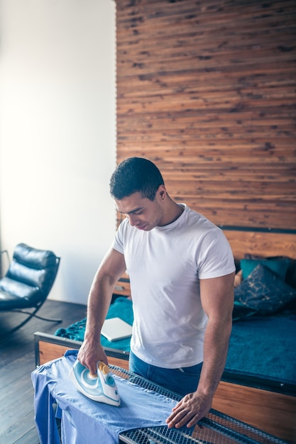 Dark-haired young man in white tshirt looking busy with ironing