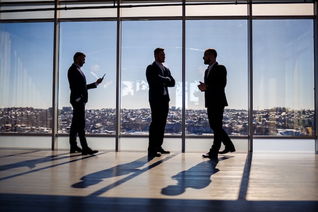 Dark male silhouettes against the background of a panoramic window. Male businessmen looking out the large window of a skyscraper overlooking the metropolis