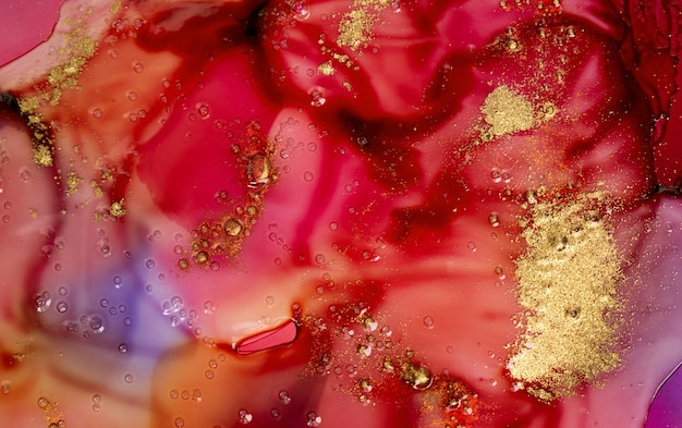 濃い赤の水彩インクは、透明な液体の下で泡と金の粒子で汚れます。