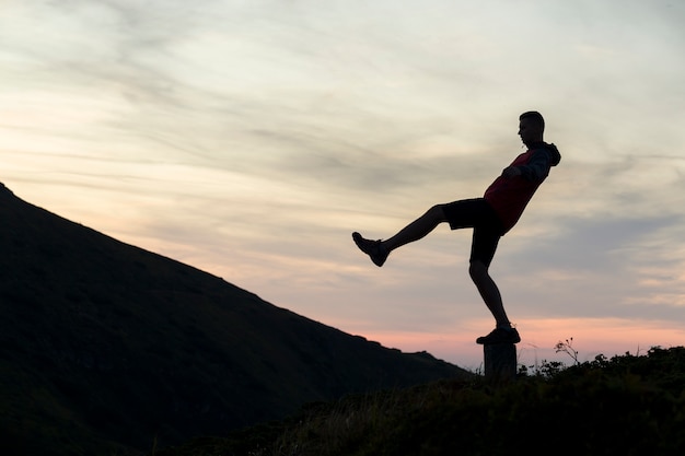 Dark silhouette of a hiker balancing on a summit stone in evening mountains.