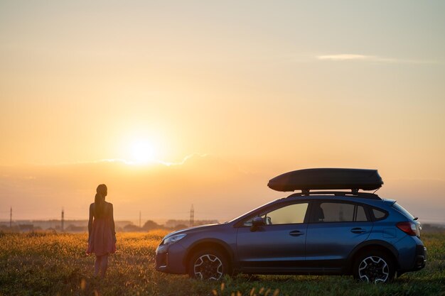 Dark silhouette of lonely woman relaxing near her car on grassy meadow enjoying view of colorful sunrise Young female driver resting during road trip beside SUV vehicle