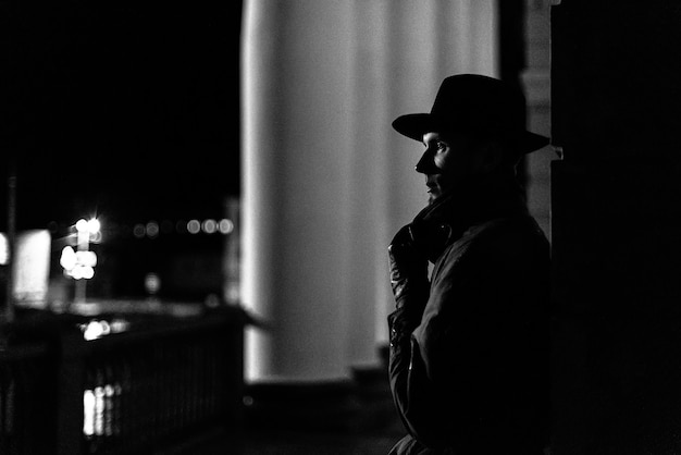 Dark silhouette of a man in a raincoat with a hat and a scar on his face at night