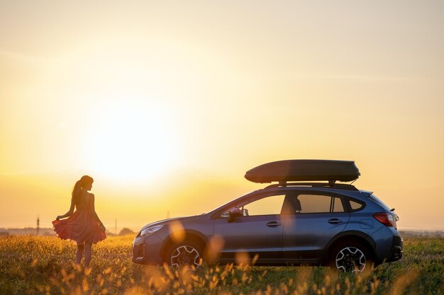 Dark silhouette of woman driver standing near her car on grassy field enjoying view of bright sunset Young female relaxing during road trip beside SUV vehicle