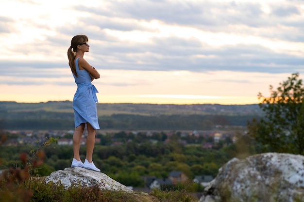 夏の屋外で夕日の景色を楽しむ石の上に立っている若い女性の暗いシルエット。