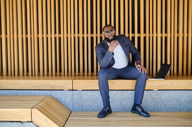 A dark-skinned businessman with a laptopsitting on the bench and working