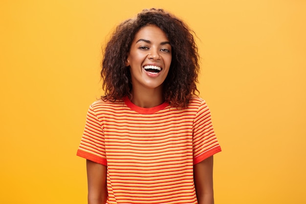 темнокожая женщина с кудрявой прической радостно смеется над оранжевой стеной
