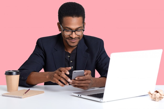 dark skinned female with modern mobile phone in hands. Handsome black man uses lap top, for working, decides checks social net work while having brake, isolated on pink