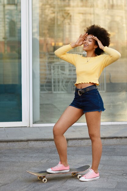 Dark-skinned girl in a yellow top and blue shorts with a skateboard against the space of the wall summer in the city