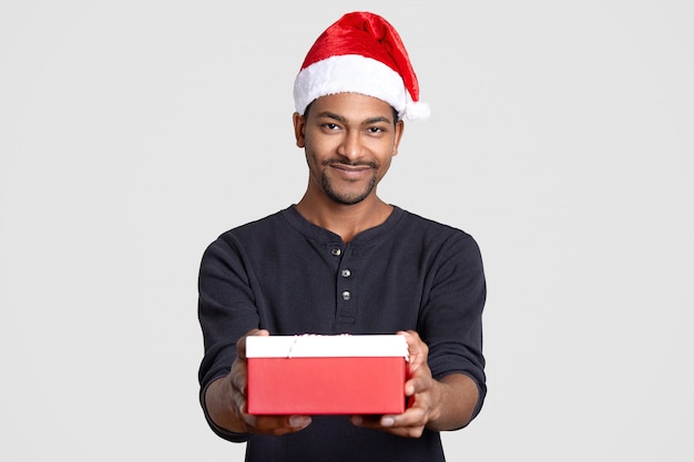 Dark skinned Santa Claus wears festive hat, casual jumper, holds present box, smiles gladfully, isolated over white