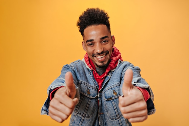 Dark skinned teen guy in denim outfit shows thumbs up on orange wall