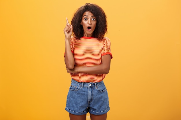 dark skinned woman staring amazed raising index finger in eureka gesture over orange wall