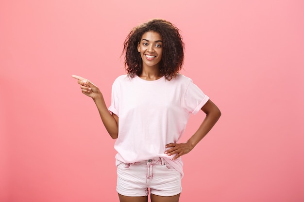 Photo dark skinned woman with curly hairstyle with hand on hip and pointing left on pink wall