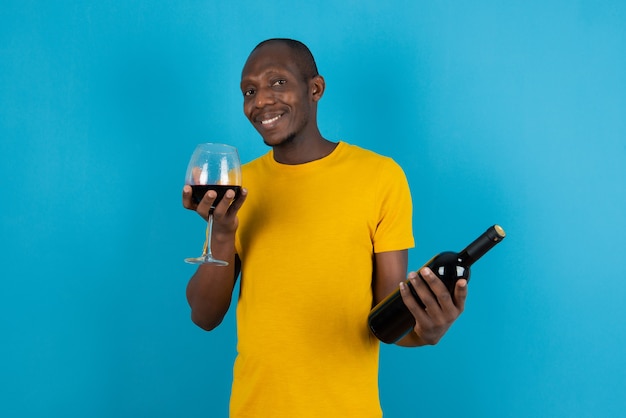 Dark-skinned young man in yellow shirt holding glass of wine on blue wall