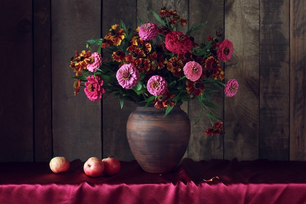 Natura morta scura con bouquet autunnale di fiori e mele in una brocca di argilla e frutta