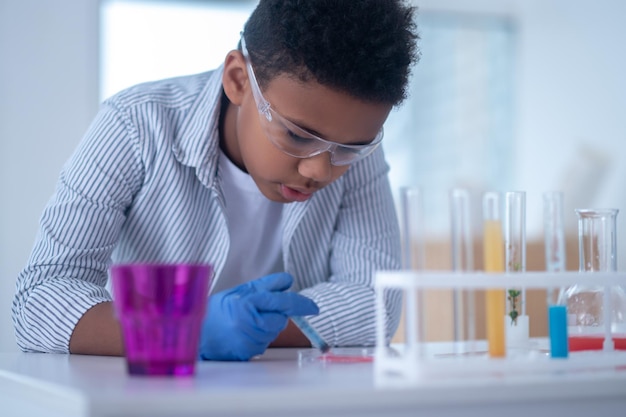 A darkhaired boy in a lab coat holding a syringe with a reagent and looking busy