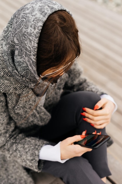 眼鏡をかけた黒髪の街の女の子が街の通りの階段に座ってスマートフォンを覗き込む